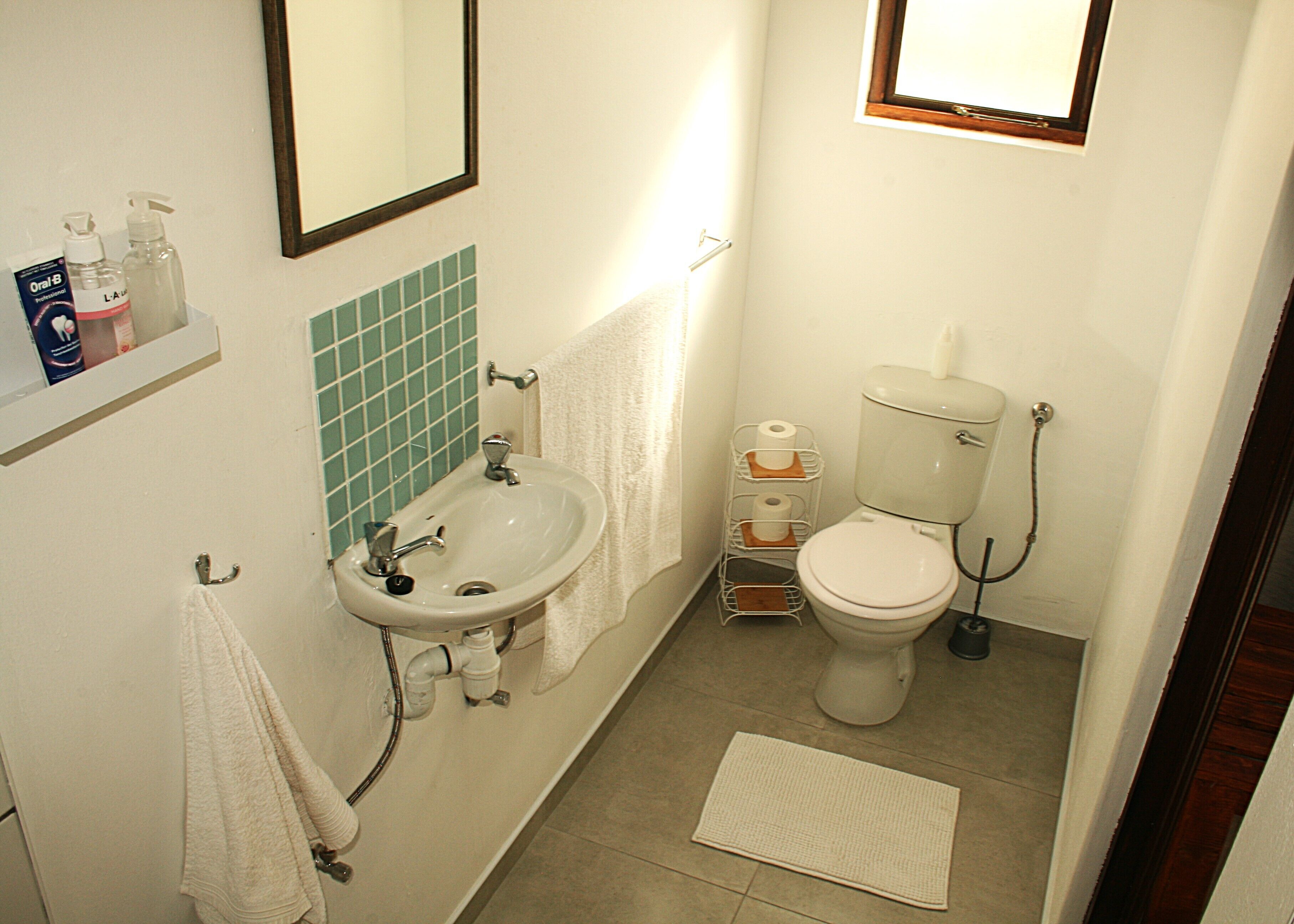 Un baño pequeño con lavamanos, inodoro y espejo. Hay artículos de aseo en la pared y un perchero para toallas.