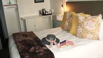 Una cama bien hecha con una manta marrón, un sombrero y un libro encima. Se ve una mesita de noche con una lámpara y una pequeña mesa con una tetera y tazas.