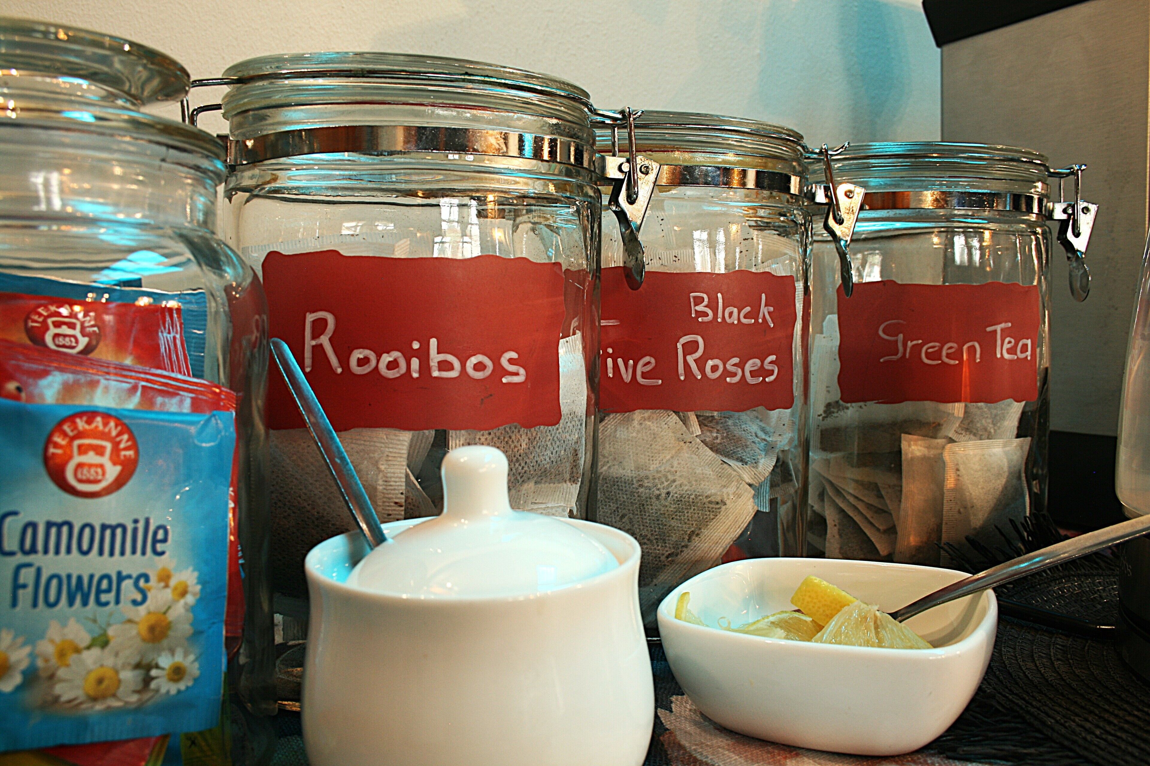 Quatre pots en verre étiquetés avec différents types de thé, une théière blanche et un bol avec une cuillère.