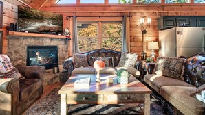 A cozy living room with a stone fireplace, leather sofas, and a large window overlooking a forest. - Smoky Mountain Ridge Retreat / Panoramic Views • Hot Tub • Game Room & Balconies (Gatlinburg)