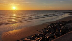 Een strand met een rotsachtige kustlijn, een pier die het water in reikt, en de zon die ondergaat aan de horizon.