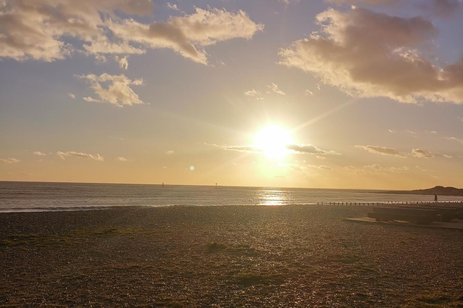 Uma praia com um píer que se estende para a água, céu claro e o sol se pondo ou nascendo.