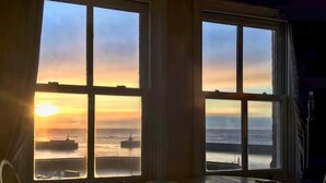 Een kamer met een groot raam met uitzicht op een zonsondergang boven de zee, een tafel en een stoel.