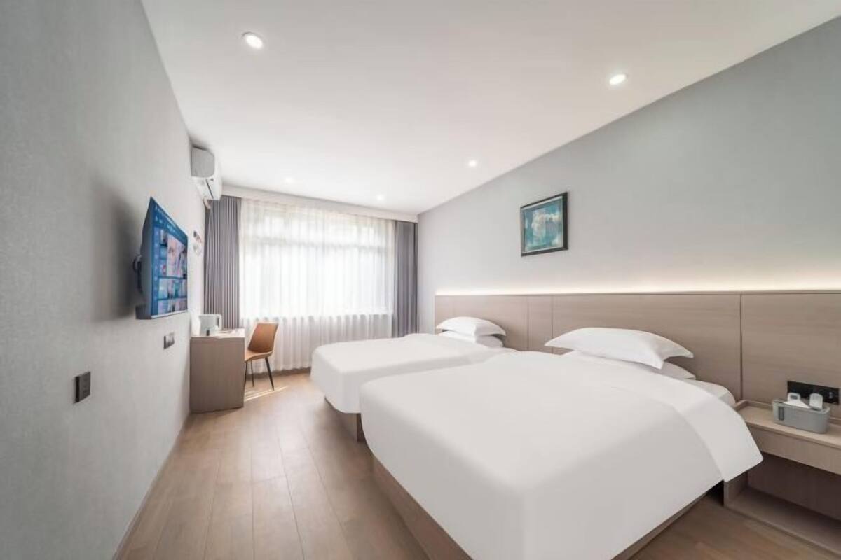 A hotel room with two beds, a desk, a chair, a window with curtains, and a painting on the wall.