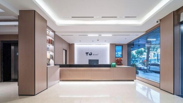 Uma área de recepção de hotel moderna com design limpo e minimalista, com balcão de recepção, uma grande janela com vista para a rua e interior bem iluminado.