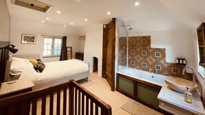 A bedroom with a bed, a wooden headboard, a bedside table, a nightstand, a window with curtains, a radiator, a wooden door, a bathroom with a bathtub, a sink, a mirror, and a tiled wall.