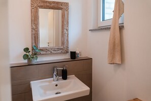 A bathroom with a white sink, a mirror with a decorative frame, a potted plant, a bottle of hand soap, and a towel hanging on a rack. - Anubis_relaxed&stay! Stylish short-term accommodation in leafy Hamburg-Niendorf (Hamburg)