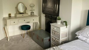 A bedroom with a white dresser, a vanity with a mirror, a green stool, a grey trunk, a fireplace, and a bed with white bedding. - Luxury Central Hale Living (Altrincham)