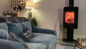 A cozy living room with a blue sofa, a decorative wall panel, a table lamp, and a wood-burning stove. - Luxury Central Hale Living (Altrincham)