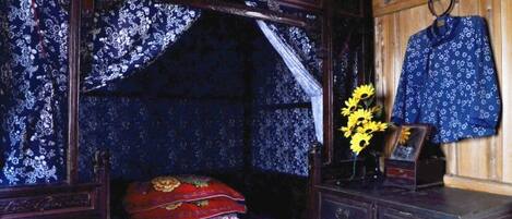 A room with a bed, a dresser, and a wall-mounted shelf with a blue patterned shirt and a vase with flowers.