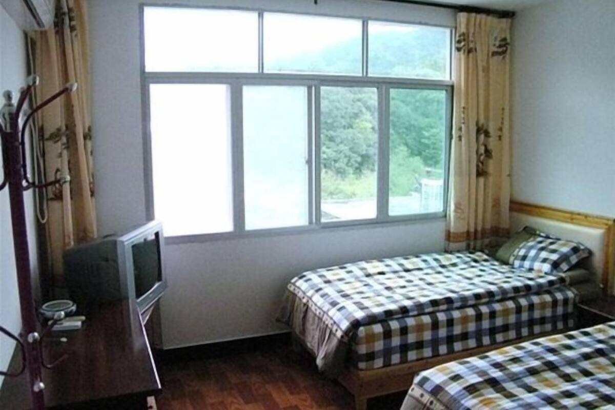 Ένα δωμάτιο με ένα μεγάλο παράθυρο, δύο κρεβάτια με καρό σεντόνια, μια ξύλινη κομοδίνα και μια τηλεόραση τοποθετημένη σε ένα έπιπλο.