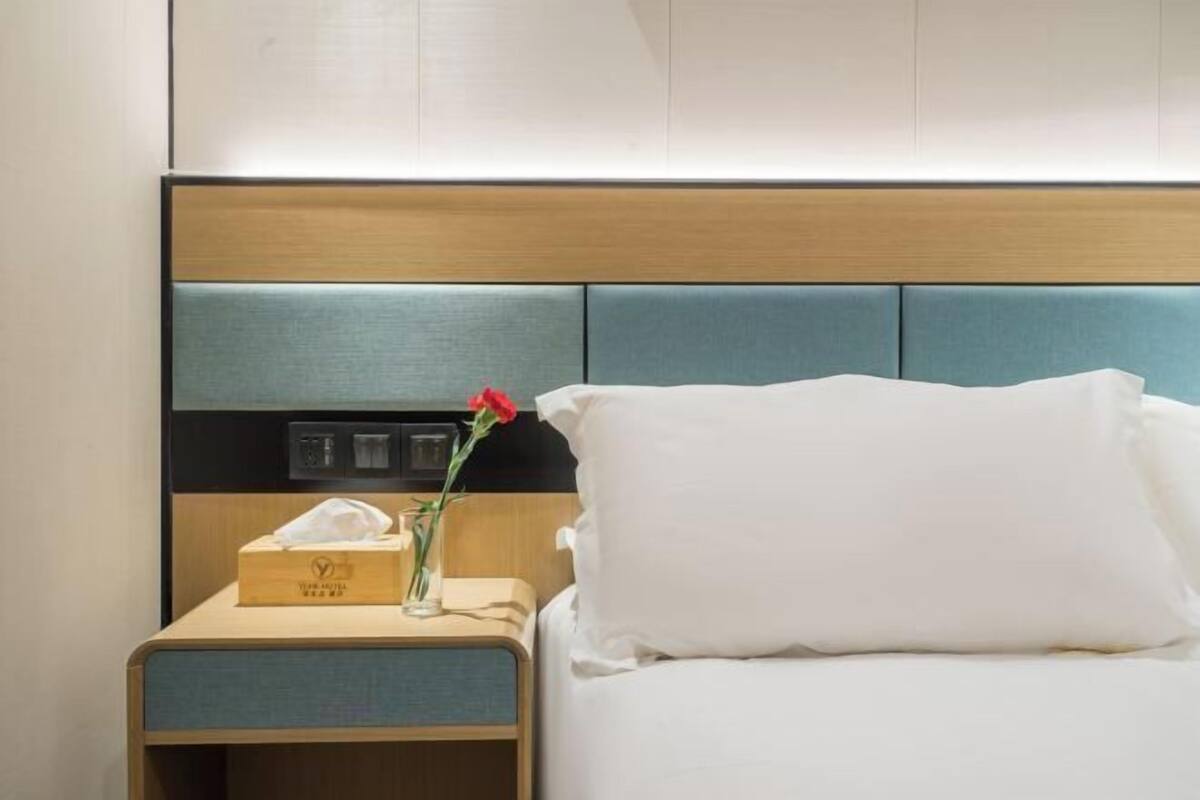 Ein Hotelzimmer mit einem Bett, einem Nachttisch mit Taschentuchbox und einer Vase mit einer Blume, sowie einem Holz-Kopfteil.