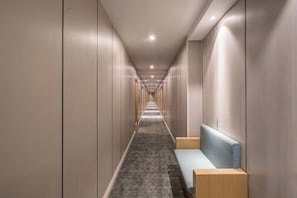 Corredor de hotel comprido e estreito, com um banco e uma mesinha de cabeceira à direita.