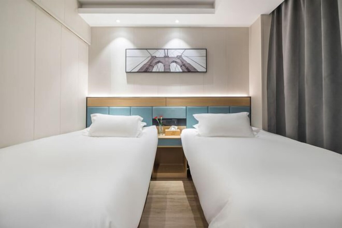 Habitación de hotel con dos camas individuales, un cabecero de madera, una mesita con un florero y un cuadro en la pared.