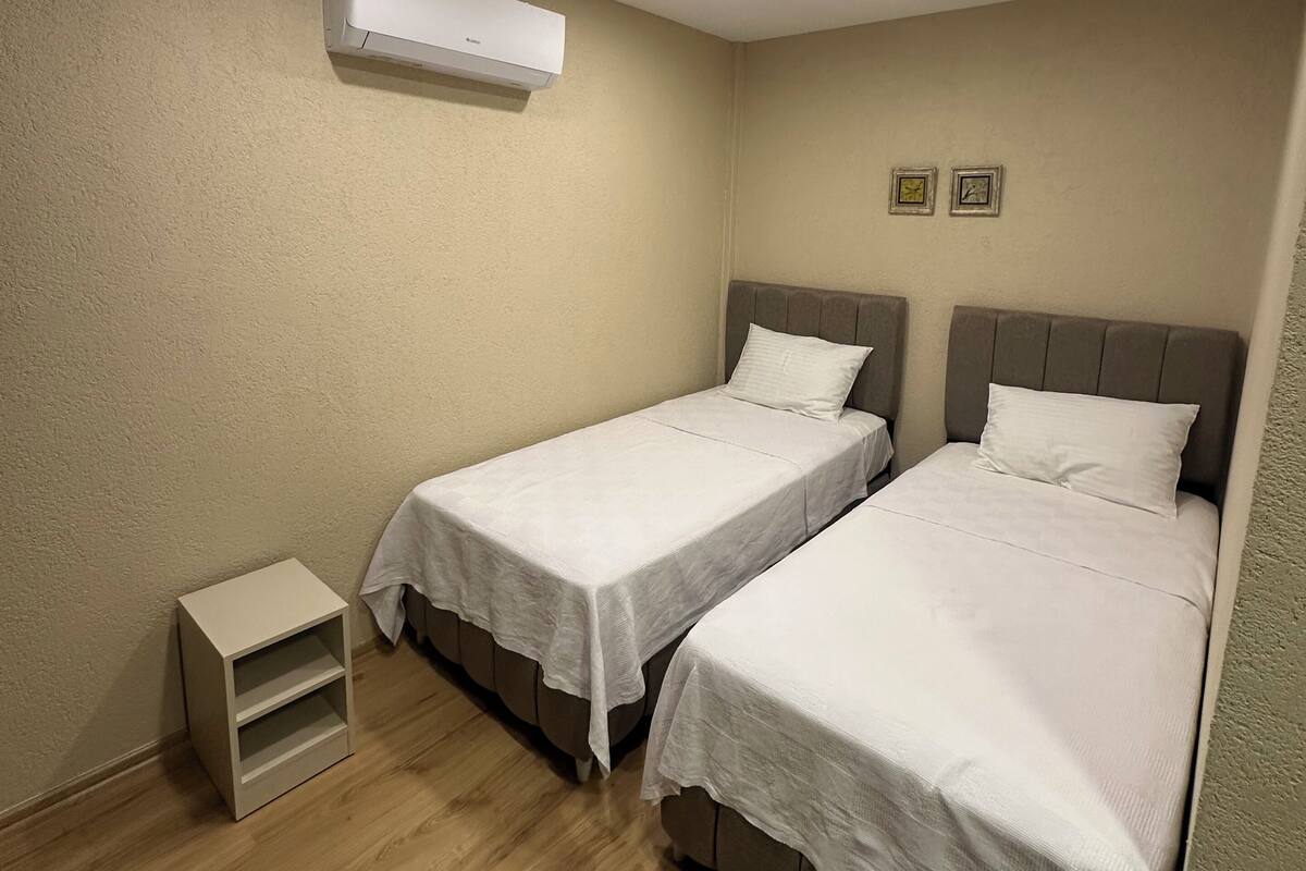 Una habitación pequeña y limpia con dos camas individuales, una mesita de noche, un equipo de aire acondicionado y una lámpara en el techo.