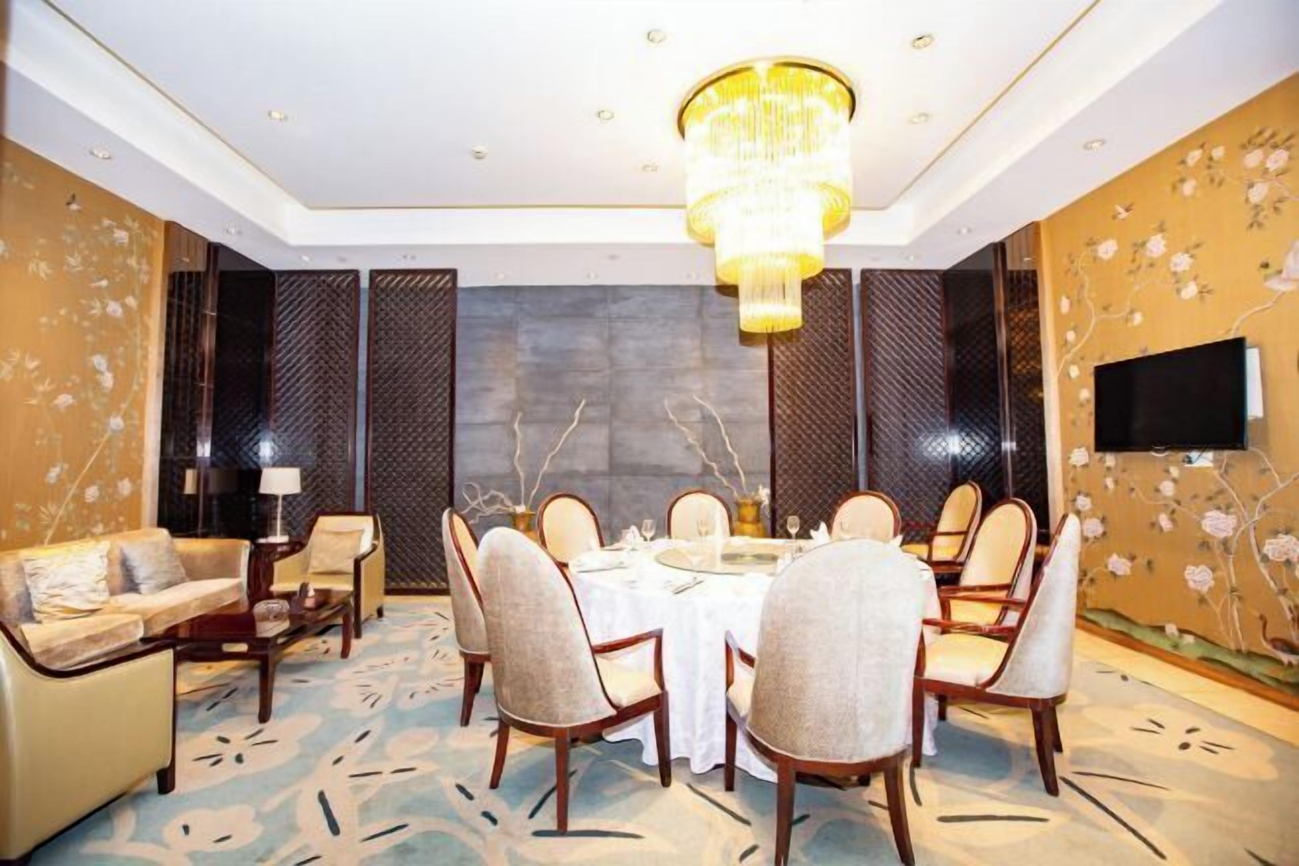 A dining room with a round table, chairs, a chandelier, a sofa, and a TV on the wall.