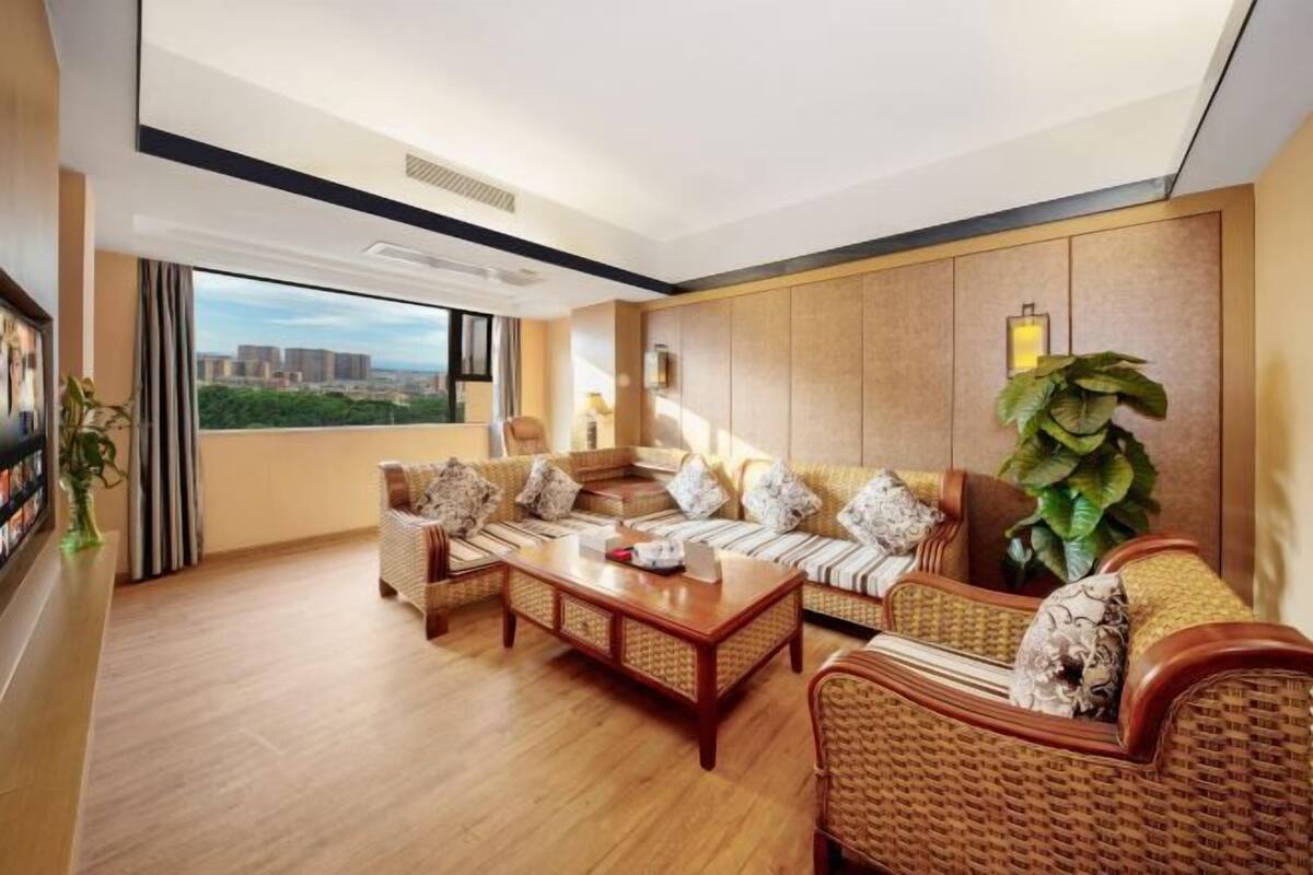 A modern living room with a wooden floor, wicker furniture, and a large window offering a city view.