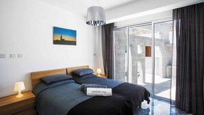 A modern bedroom with a large bed, a nightstand with a lamp, a painting on the wall, and a view of the outside through a glass door. - Villa Cerulean Paphos (Paphos)