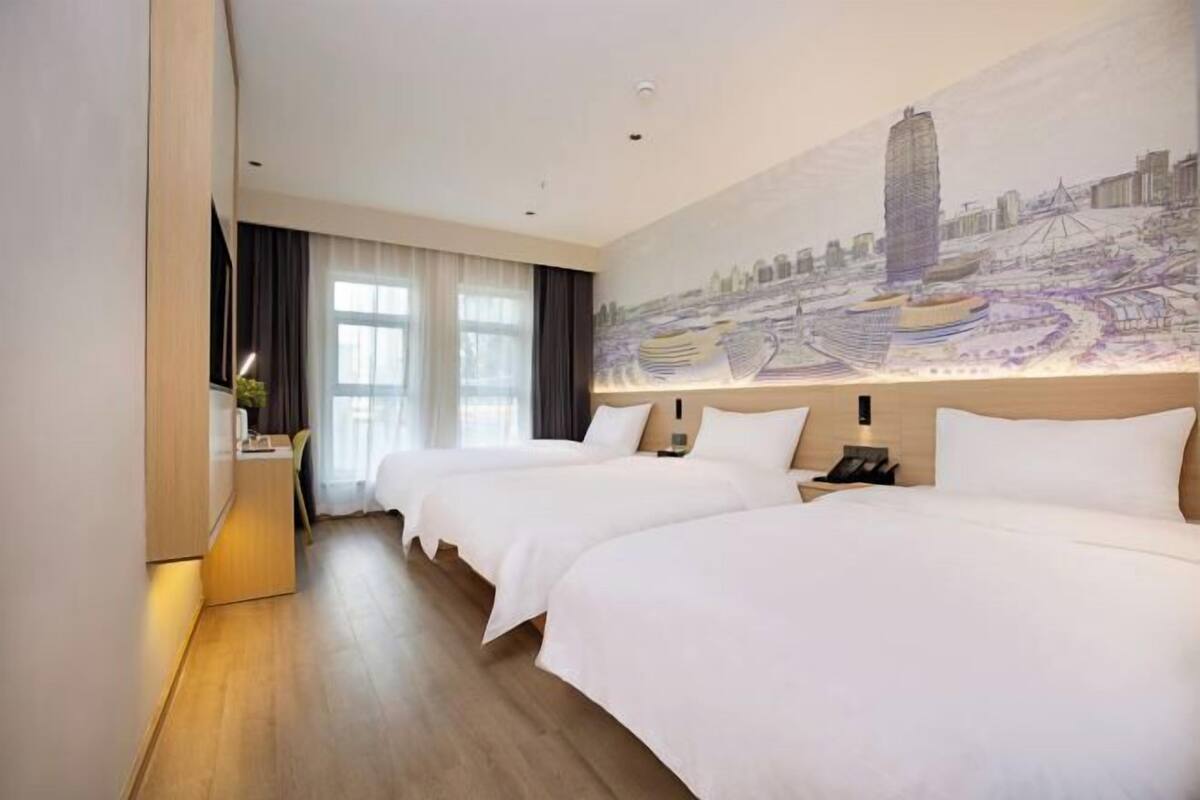 Habitación de hotel con dos camas, un mural de paisaje urbano, un escritorio con lámpara y vista a la ciudad.