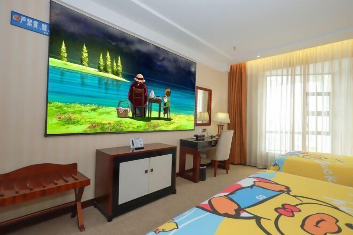A hotel room with a large TV displaying a nature scene, a desk with a chair, a wooden bench, and a bed with cartoon-themed bedding.