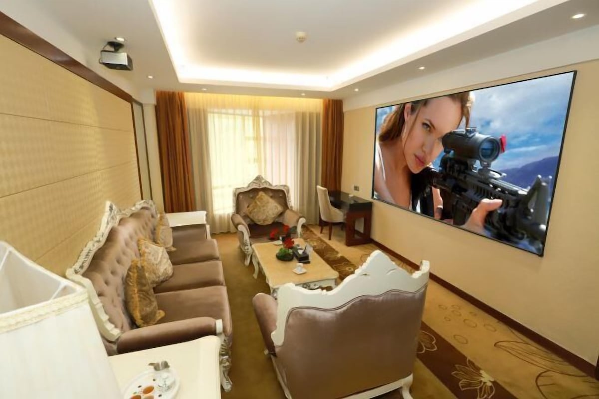 A hotel room with a large flat-screen TV, a sofa, a coffee table, and a dining area.