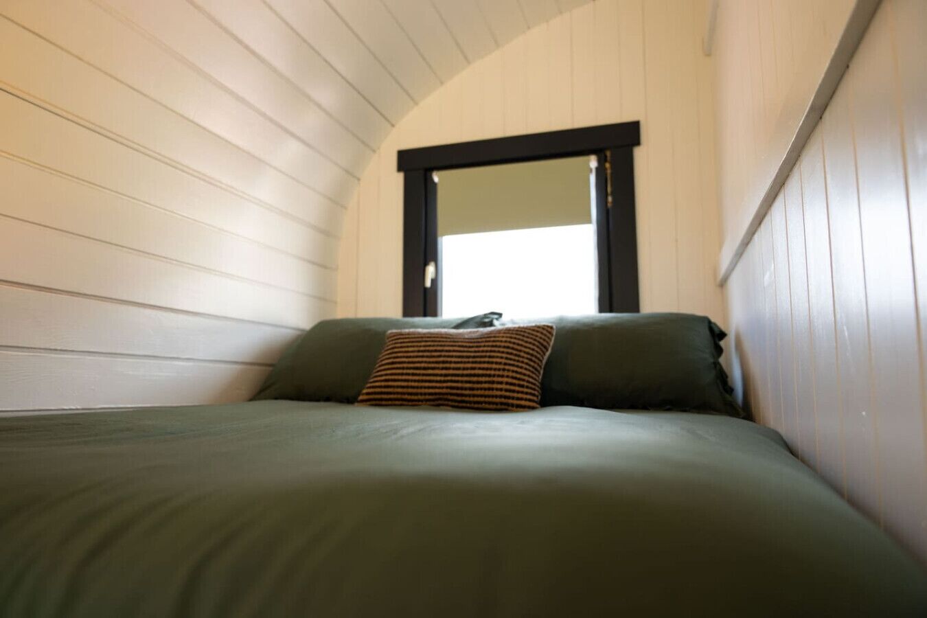 A single bed with a green mattress and two pillows, one striped, in a room with white paneled walls and a black-framed window.