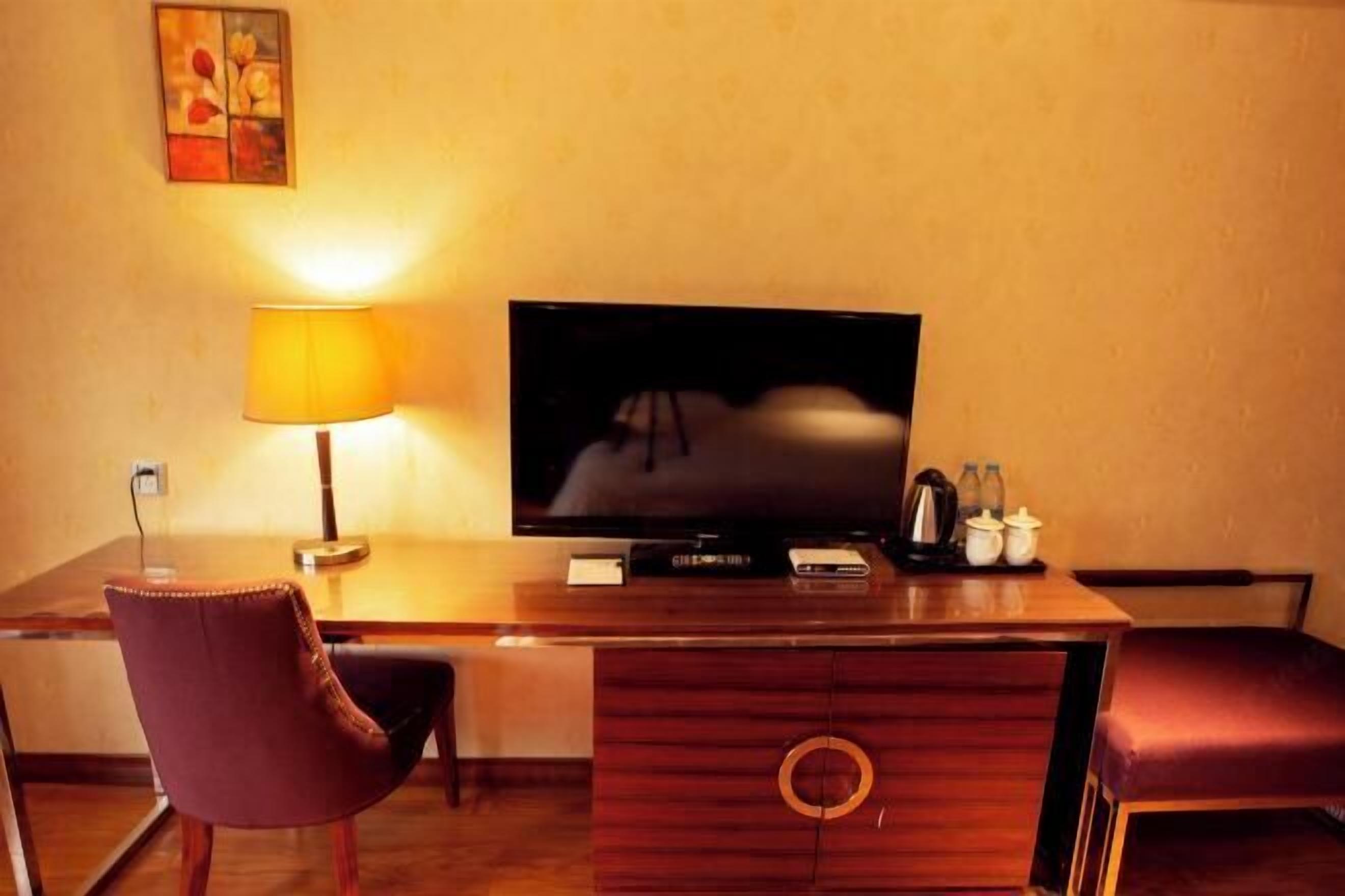 A hotel room with a wooden desk, a television, a lamp, a chair, and a small table with a teapot and cups.