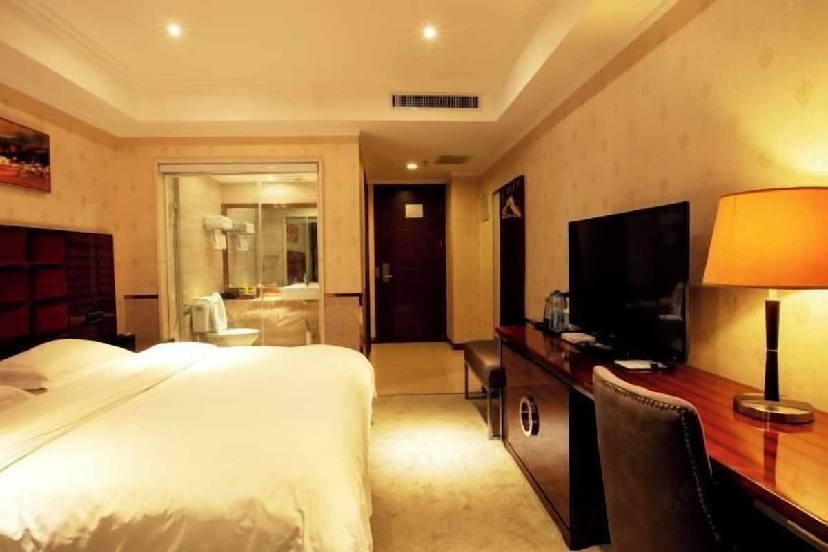 A hotel room with a large bed, a desk with a lamp, a television, and a bathroom with a shower and toilet.