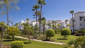 A well-maintained garden with palm trees, neatly trimmed bushes, and a clear pathway leading to a row of white buildings with balconies.