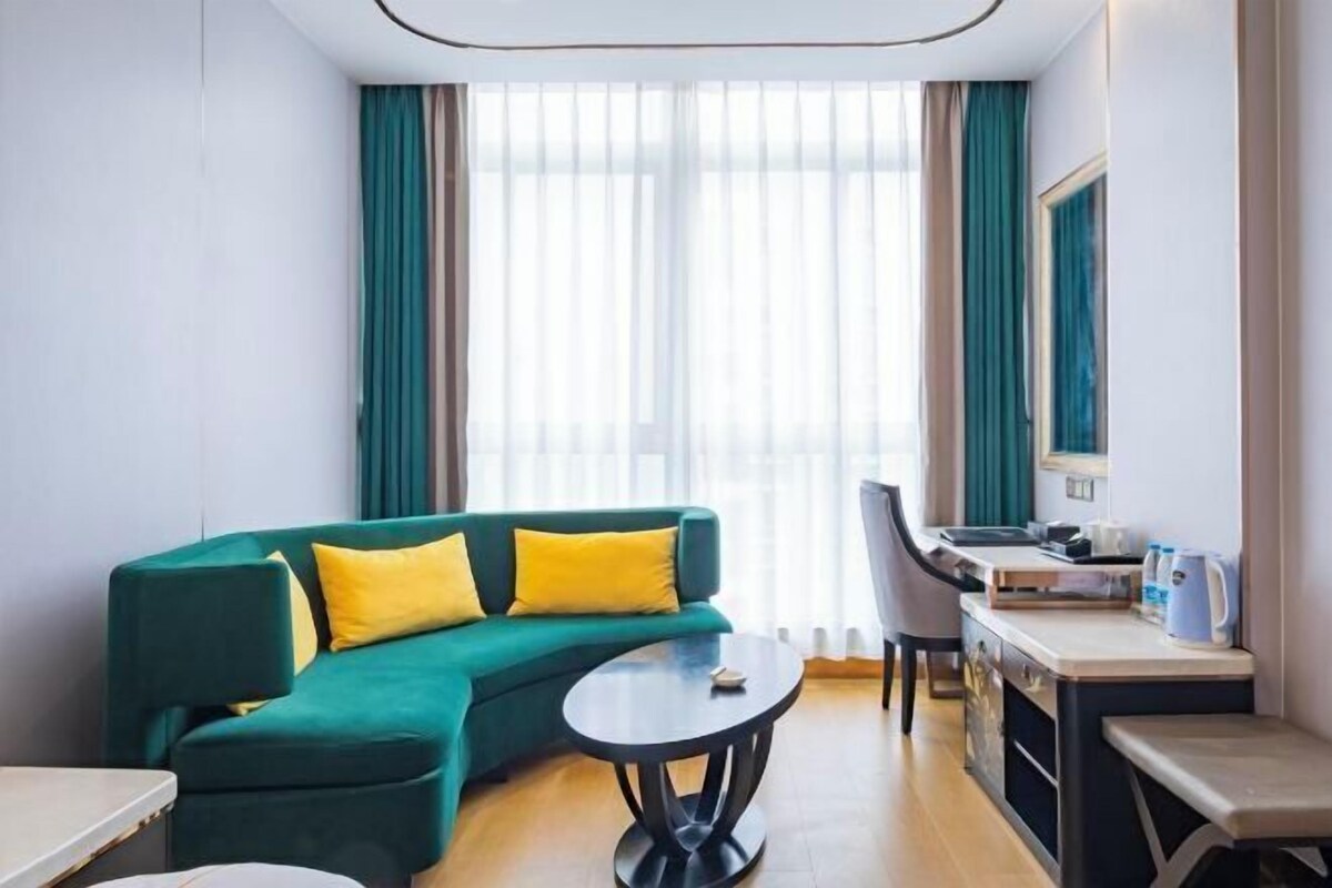 A modern hotel room with a green sofa, a round table, a desk, and a large window with curtains.