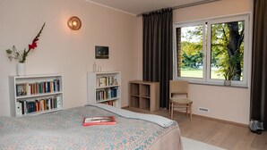 Ein Schlafzimmer mit einem Bett, zwei Bücherregalen, einem Sessel, einem Fenster mit Vorhängen und einem kleinen Tisch.