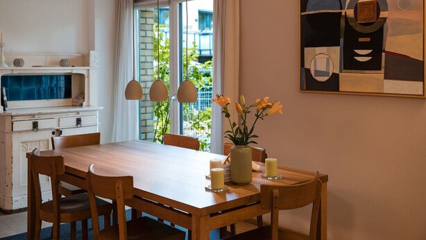 Ein Essbereich mit Holztisch, Stühlen, einer Vase mit Blumen und Kerzen. Ein Fenster mit Vorhängen, eine Mikrowelle und ein Gemälde an der Wand sind ebenfalls vorhanden.