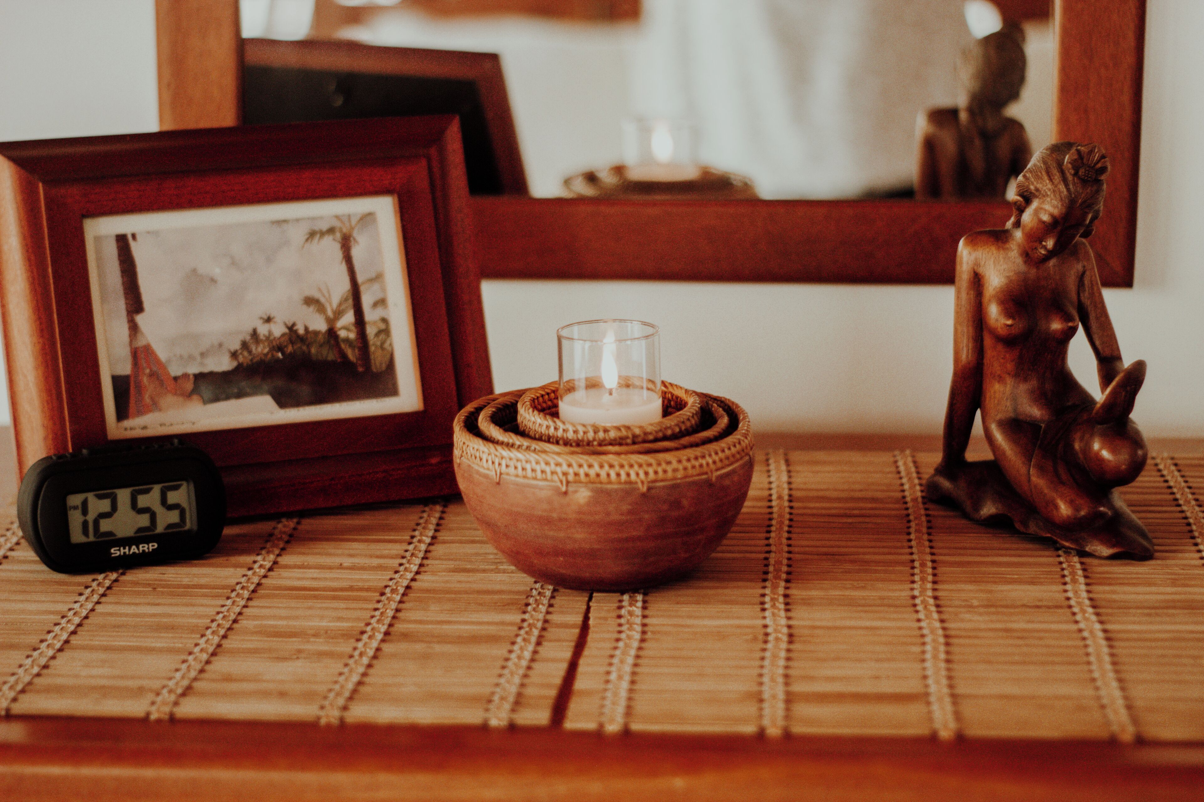 A wooden table with a framed picture, a small clock, a candle in a woven bowl, and a statue.