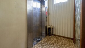 Een hal met een metalen lift, een kleine prullenbak en een raam met jaloezieën.