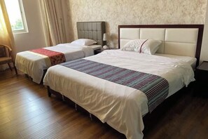 Een hotelkamer met twee bedden, houten vloeren, een groot raam met gordijnen en een nachtkastje met een lamp.