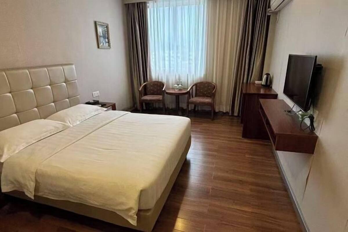 Une chambre d’hôtel avec un grand lit, une télévision, une petite table avec des chaises et une fenêtre avec des rideaux.