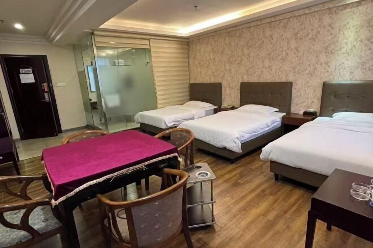 Une chambre d’hôtel avec deux lits, une table à manger recouverte d’une nappe rose, des chaises en bois, une petite table avec une lampe et une cloison en verre.