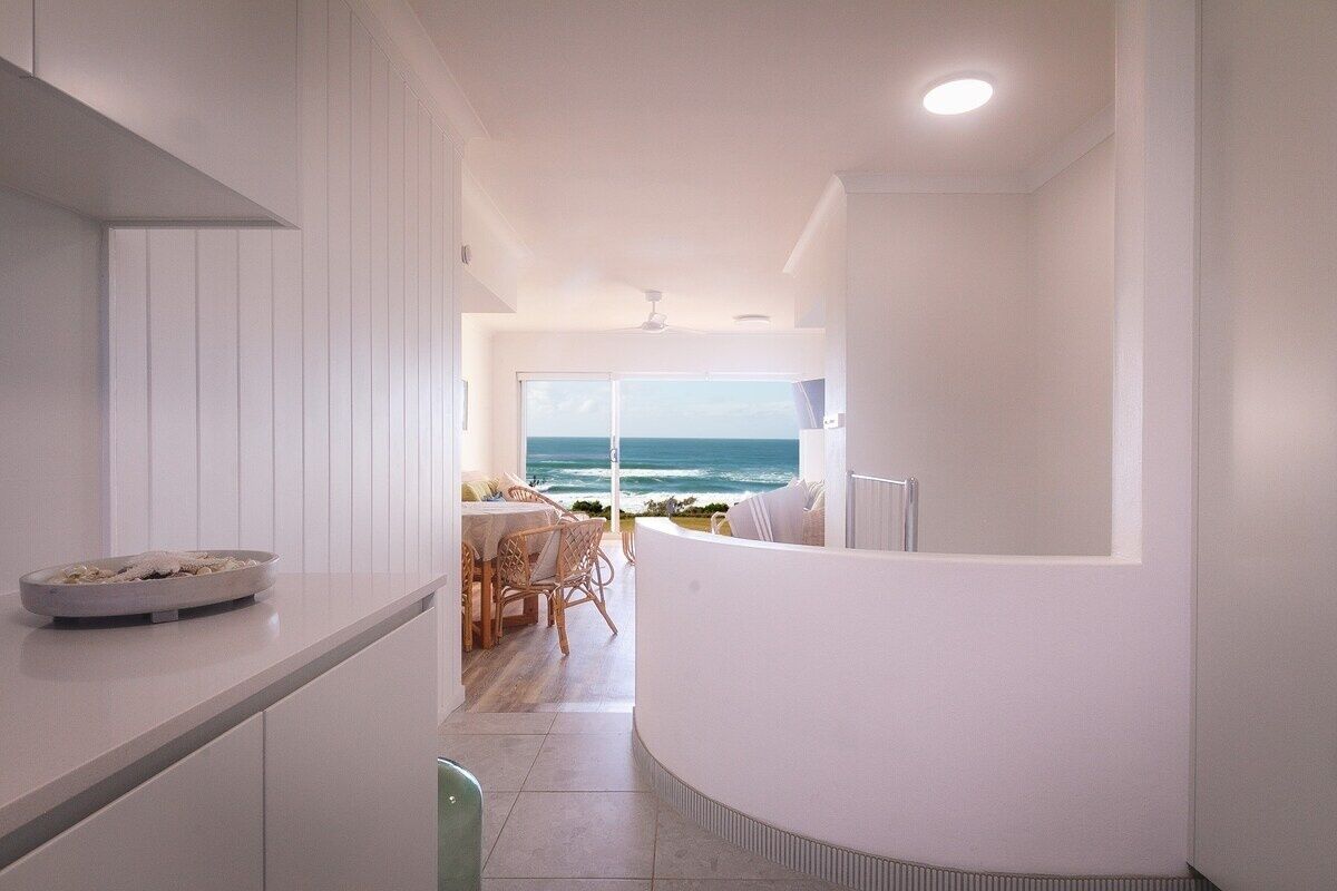 Cocina moderna con vista al océano, comedor con mesa y sillas, y decoración en tonos blancos.