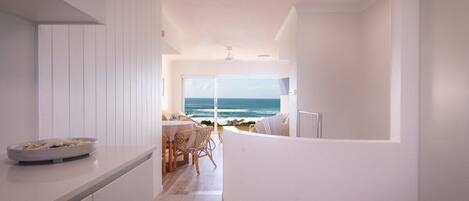 Cocina moderna con vista al océano, comedor con mesa y sillas, y decoración en tonos blancos.