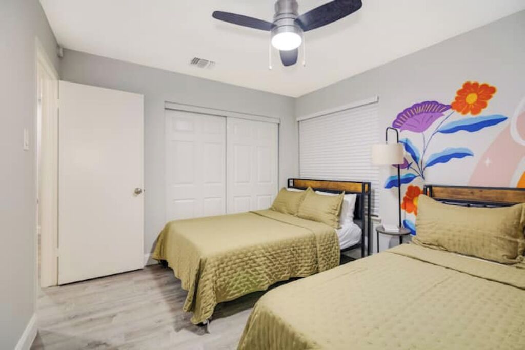 A bedroom with two beds, a ceiling fan, a door, and a wall with a floral mural.