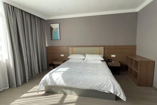 Une chambre d’hôtel avec un lit, des tables de chevet, une petite table, une tête de lit en bois, un tableau au mur et une grande fenêtre avec des rideaux.