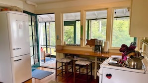 Eine kleine Küche mit Kühlschrank, Herd und Kaffeemaschine. Es gibt Hocker und ein Fenster mit Blick auf Bäume.