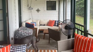 Eine gemütliche Veranda mit weißer Backsteinwand, einem Korbstuhl, einer Couch mit gemusterten Kissen und einem großen Ventilator.