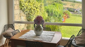 Un coin repas avec une table et des chaises, un vase rempli de fleurs et une vue sur la verdure et les collines, visible à travers une grande fenêtre en arc de cercle.