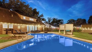 A swimming pool with lounge chairs and string lights, surrounded by a brick building with a thatched roof. - The Whimsy Retreat-amazing pool-5Brs-houston prime location Beltway 8 & US 69 (Houston)