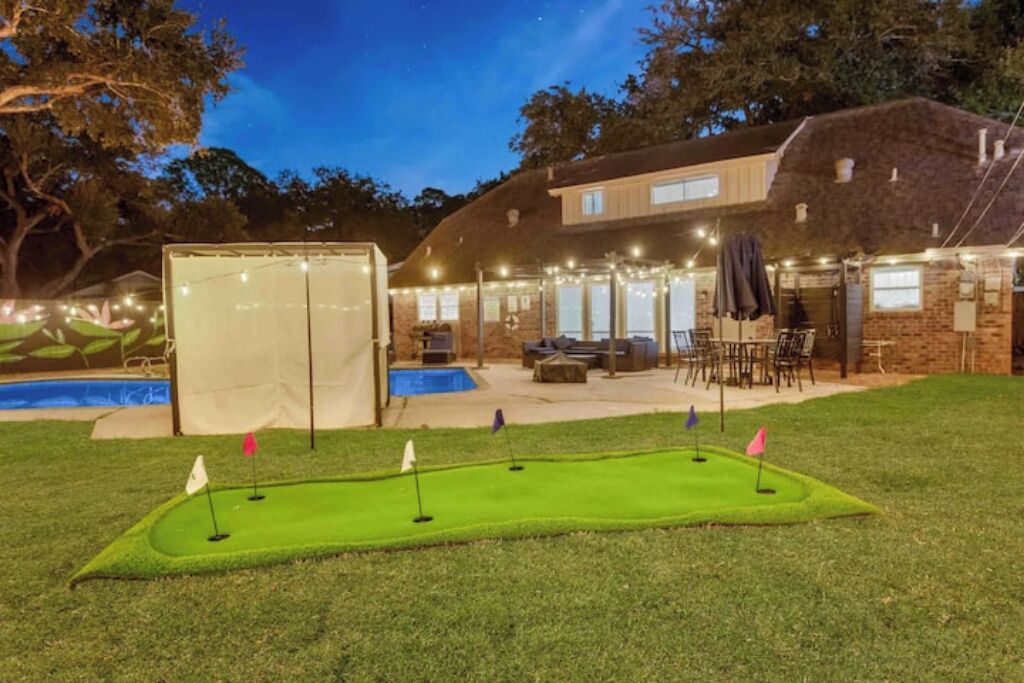 A backyard with a putting green, a swimming pool, and a covered patio area.