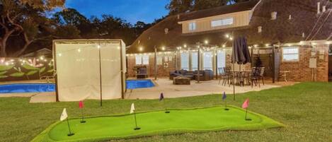 A backyard with a putting green, a swimming pool, and a covered patio area.