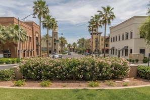 A street view with palm trees, a flower bed, and buildings with a brick facade and a beige building with a flat roof. - Desert Oasis Buckeye Gameroom Pool Golf (Buckeye)