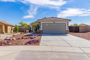 A residential house with a garage, a driveway, and a landscaped garden with rocks and purple flowers. - Desert Oasis Buckeye Gameroom Pool Golf (Buckeye)