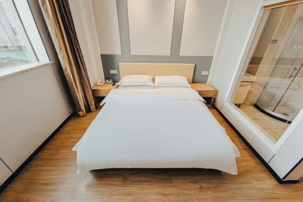 A modern hotel room with a large bed, wooden flooring, a nightstand, and a window with curtains.